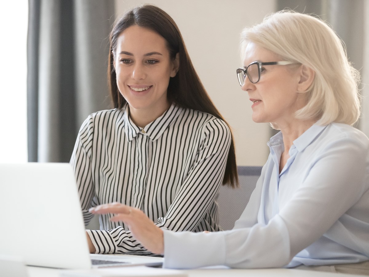 Two women sitting on a table working with a laptop. One is coaching the other on how to achieve her goals.